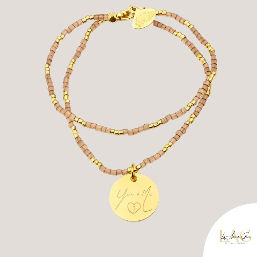 Gold-tone beaded bracelet with small gold spacers and a round pendant engraved 'You + Me' with a heart symbol, on a light beige background.