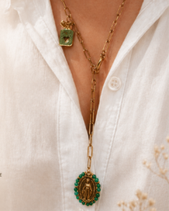 Gold multi-layered necklace with a green-beaded oval pendant featuring a religious figure, worn over a white blouse.